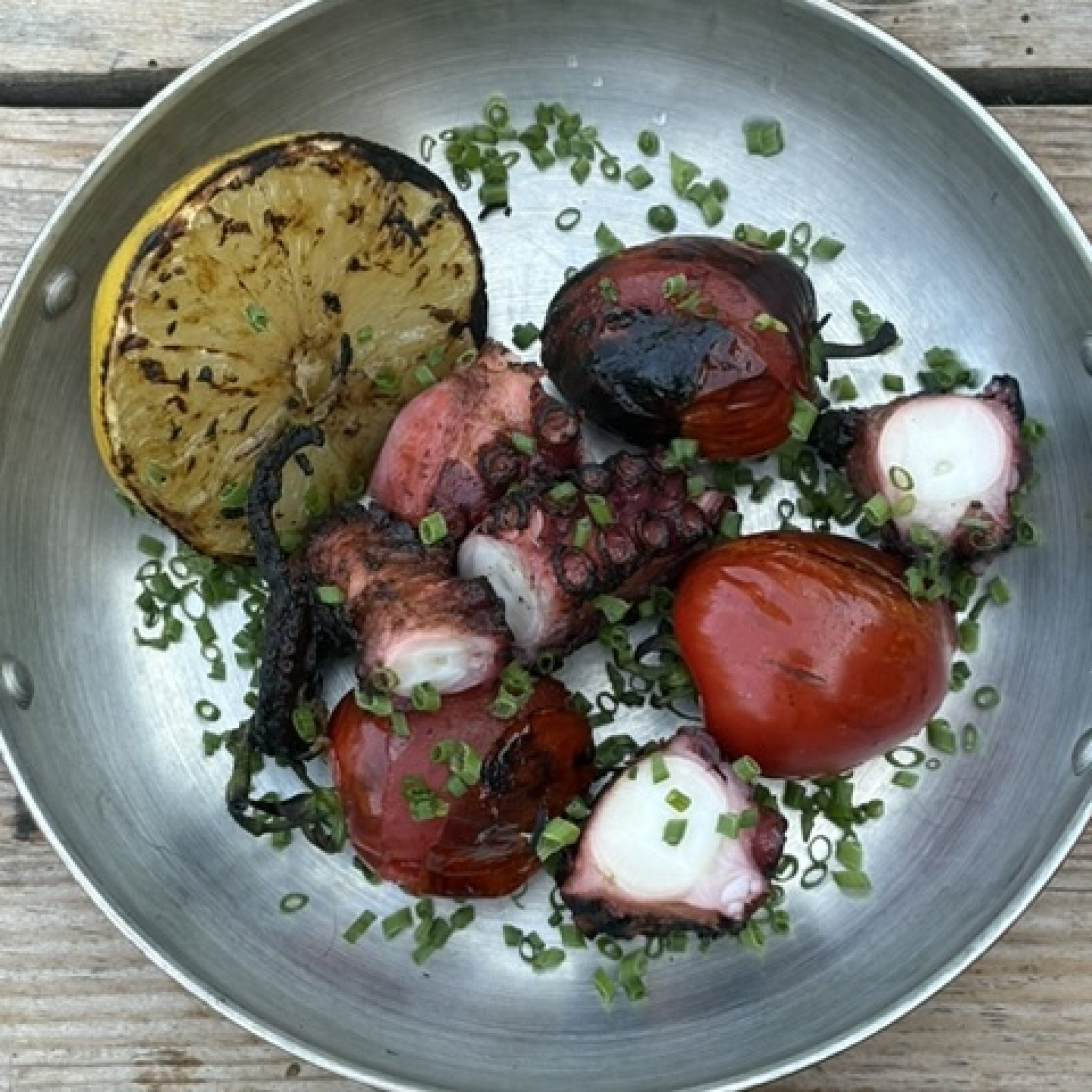 Grillad pulpo med brända tentakler i en stålpanna, tillsammans med grillad citronhalva, tomater och gräslök
