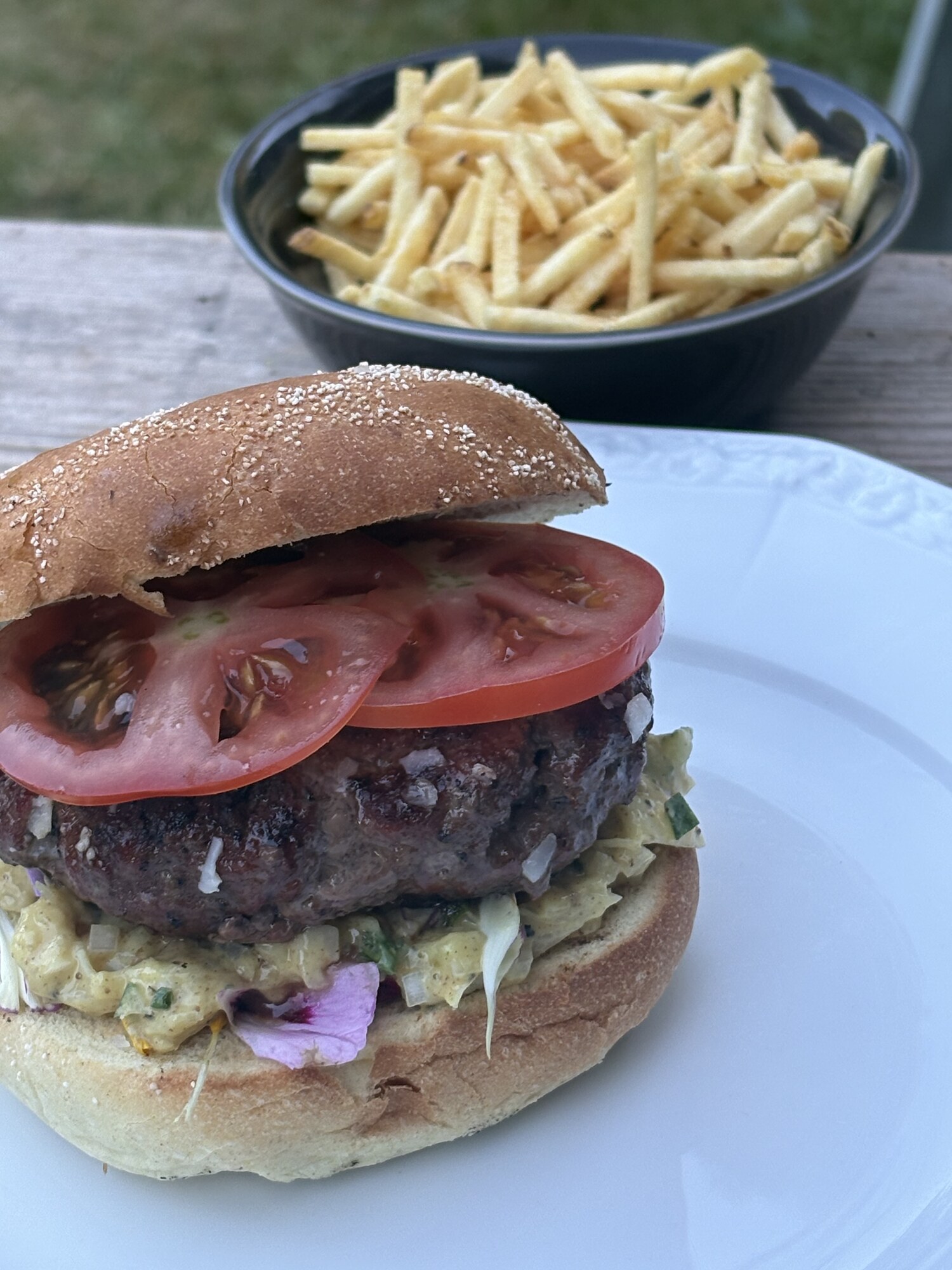 Hamburgare på pata negra-färs i sesambröd med tomatskivor och kålsallad, med en skål tunna pommes bakom