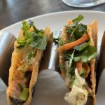 Två friterade tacoskal i stålhållare, fyllda med kött, strimlad morot, kål och rikligt med koriander