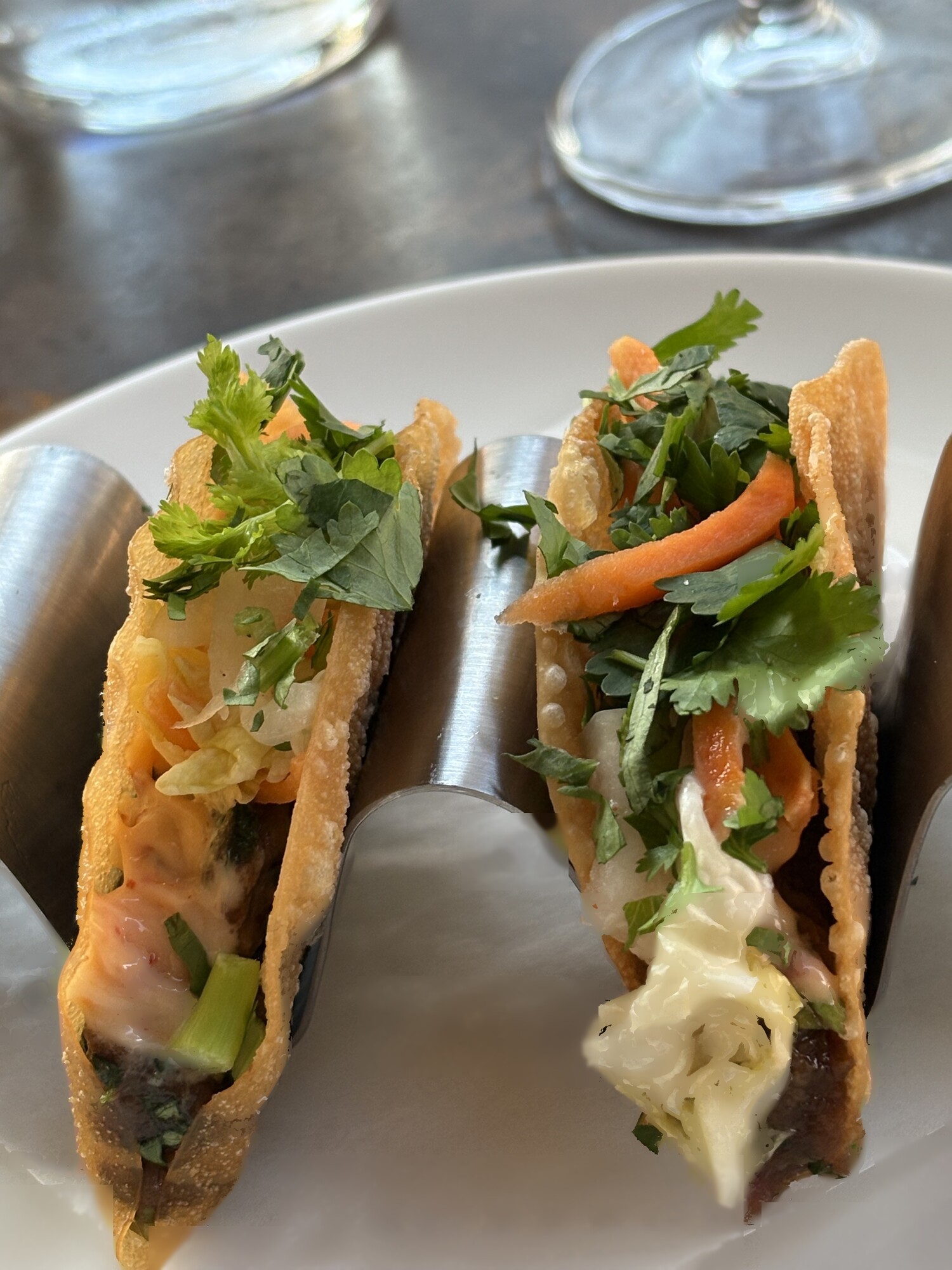Två friterade tacoskal i stålhållare, fyllda med kött, strimlad morot, kål och rikligt med koriander