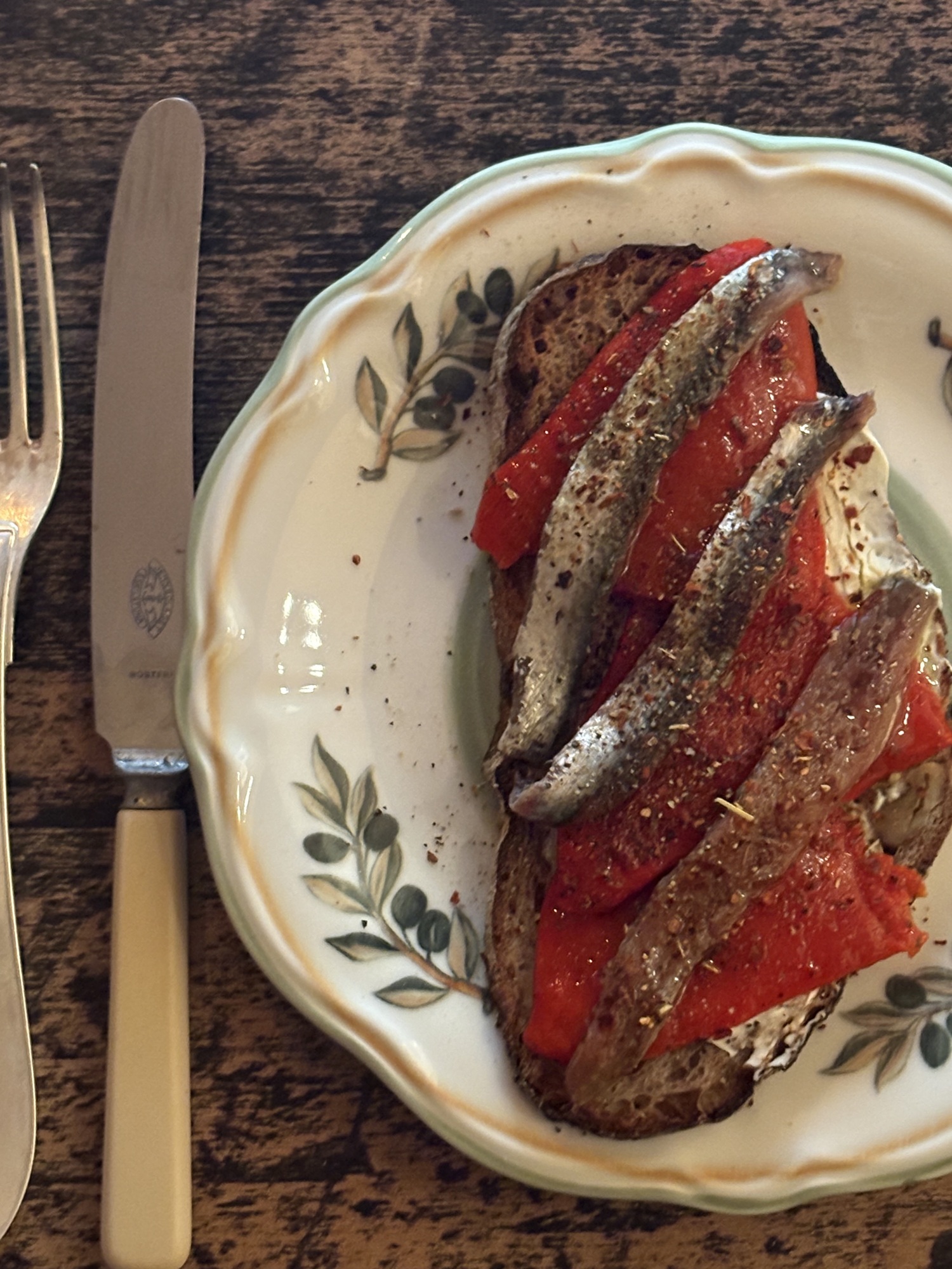 Grillad brödskiva täckt med rostad piquillopaprika och ansjovisfiléer på en olivmönstrad tallrik, med kniv och gaffel bredvid