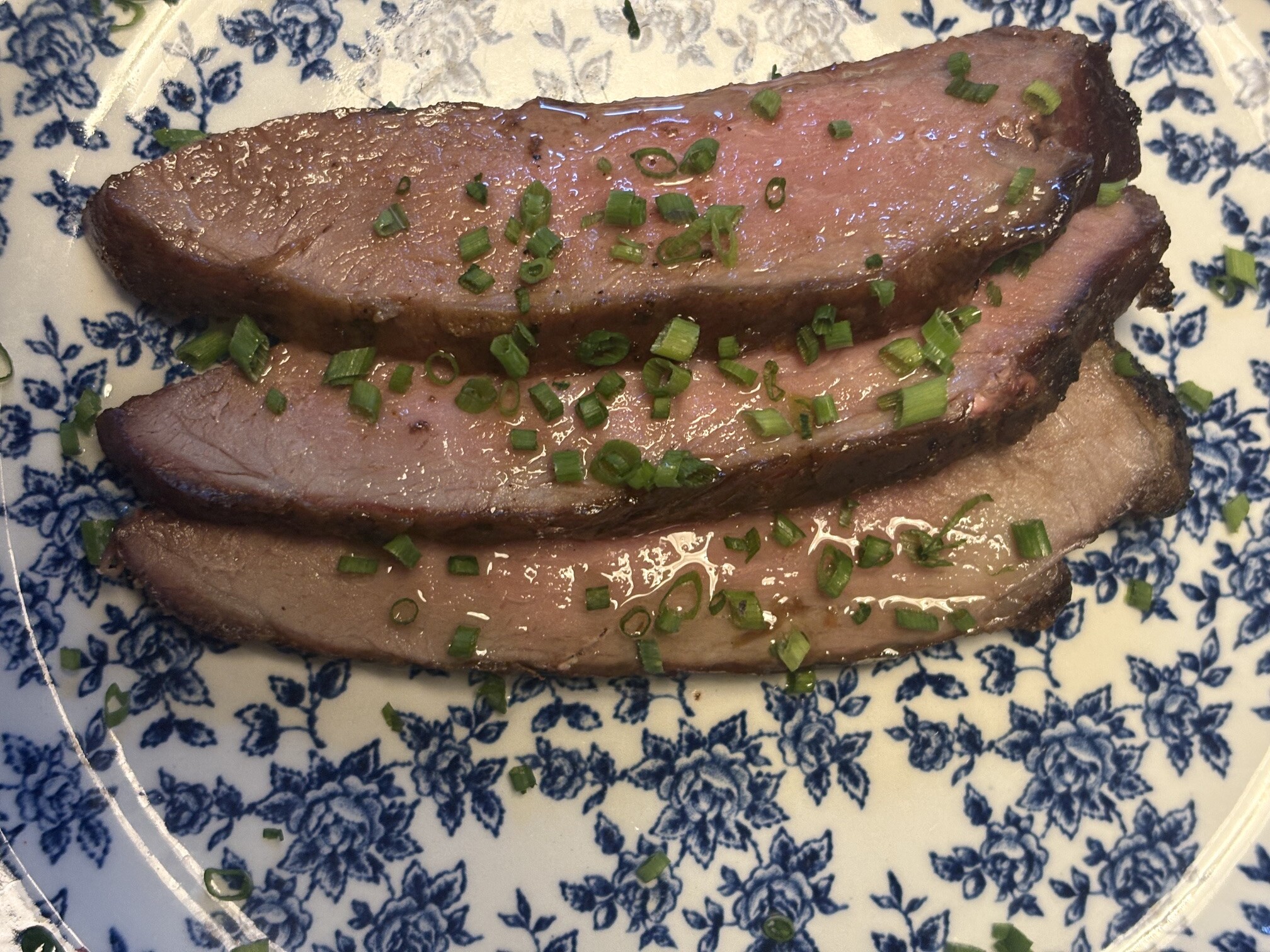 Tre skivor grillad presa pata negra med rosa kärna och blank sky, strödda med hackad salladslök på blåblommig tallrik