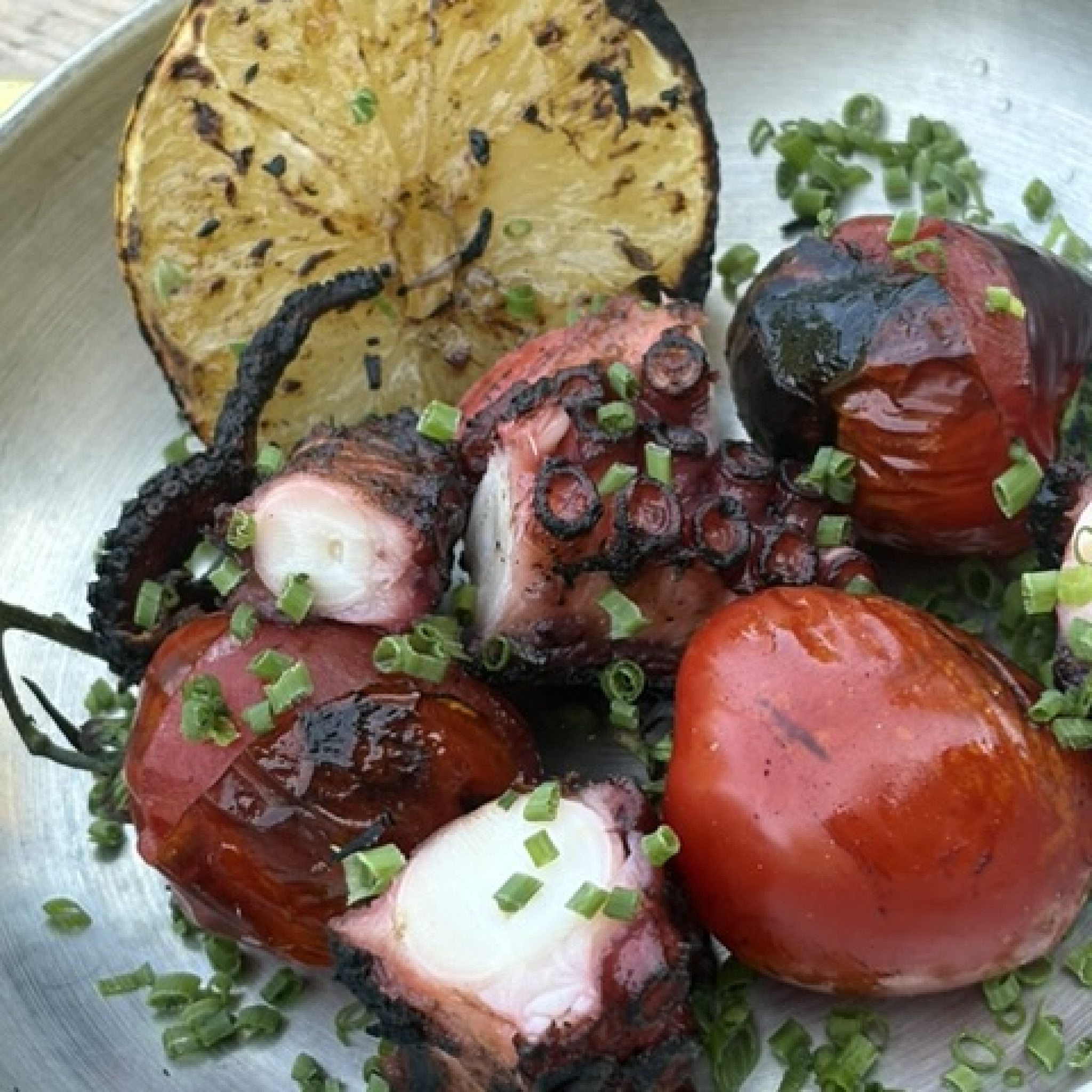 Närbild på grillad pulpo med brända sugkoppar i stålpanna, med grillad citronhalva, tomater och gräslök