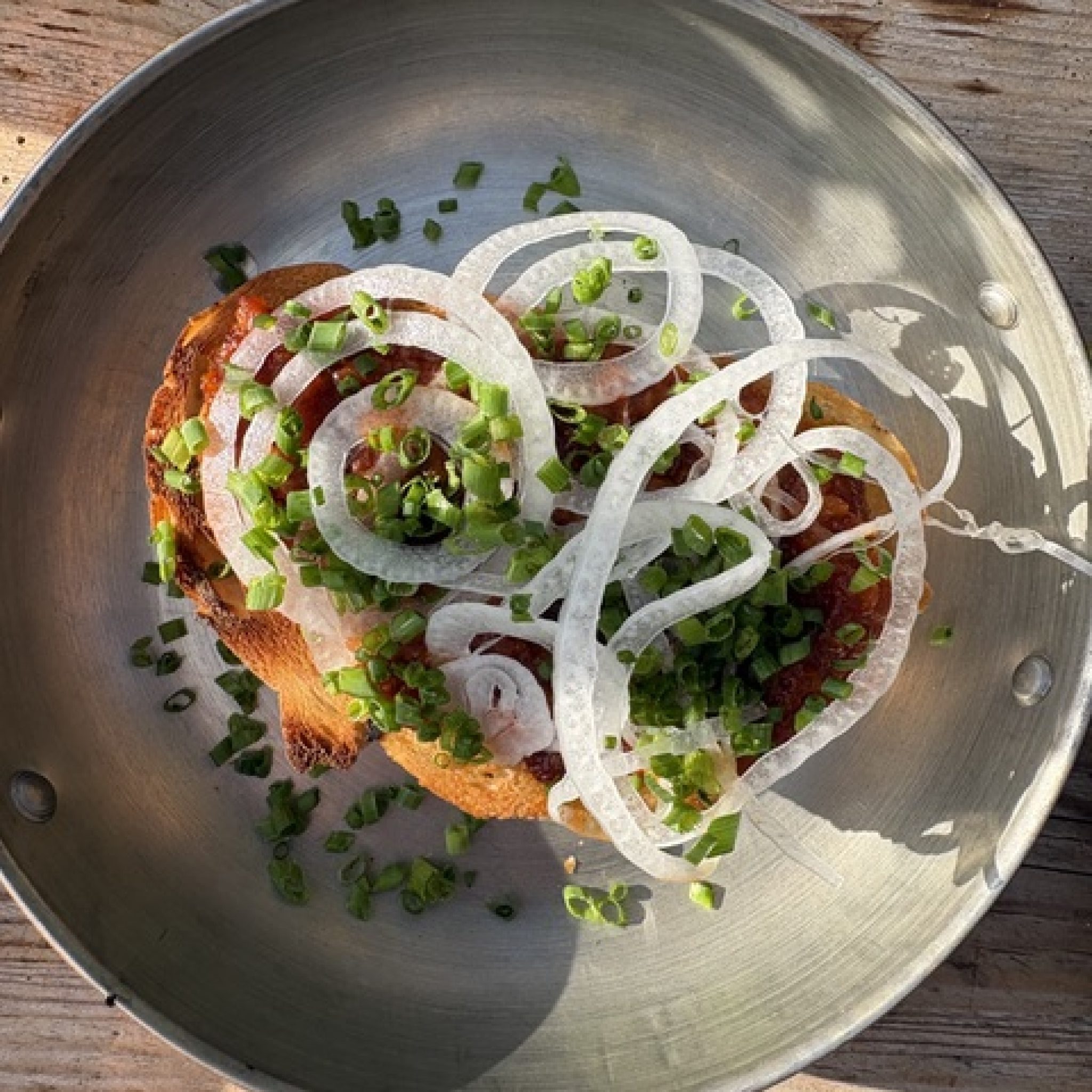 Grillat surdegsbröd med sobrasada i stålpanna, täckt av papperstunna lökringar och gräslök
