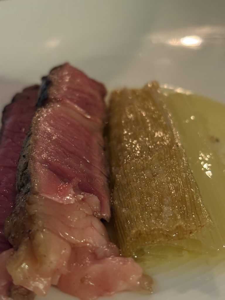 Skivad grillad biff med rosa kärna bredvid en bakad purjolökshalva med flingsalt på vit tallrik