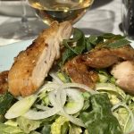 Krispigt stekt kycklinglår på sallad av spetskål, salladslök och örter, med ett glas vitt vin i bakgrunden
