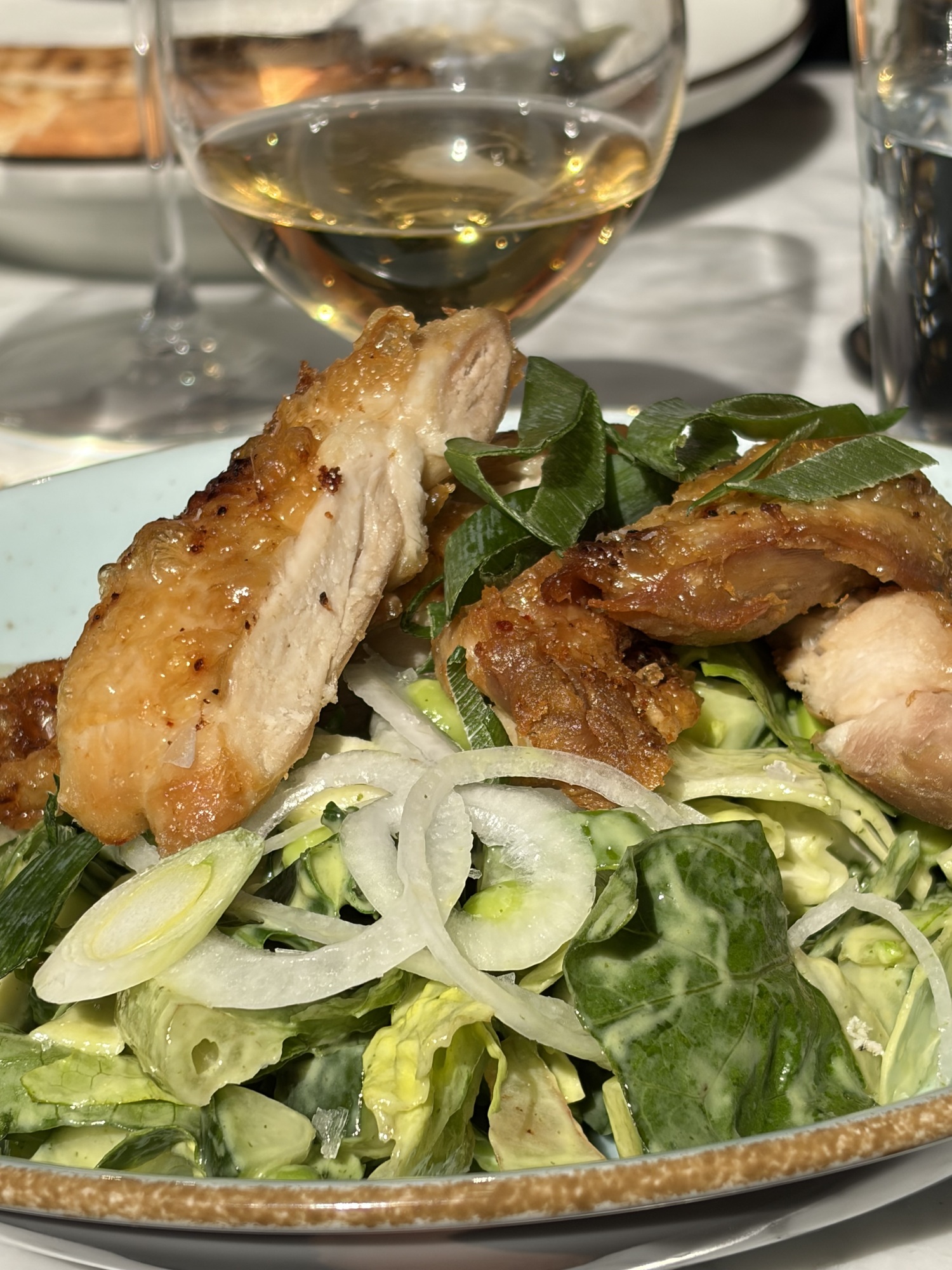Krispigt stekt kycklinglår på sallad av spetskål, salladslök och örter, med ett glas vitt vin i bakgrunden