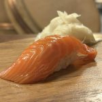 Nigiri med tjock laxskiva över risbädd på träbricka, med syltad ingefära bakom