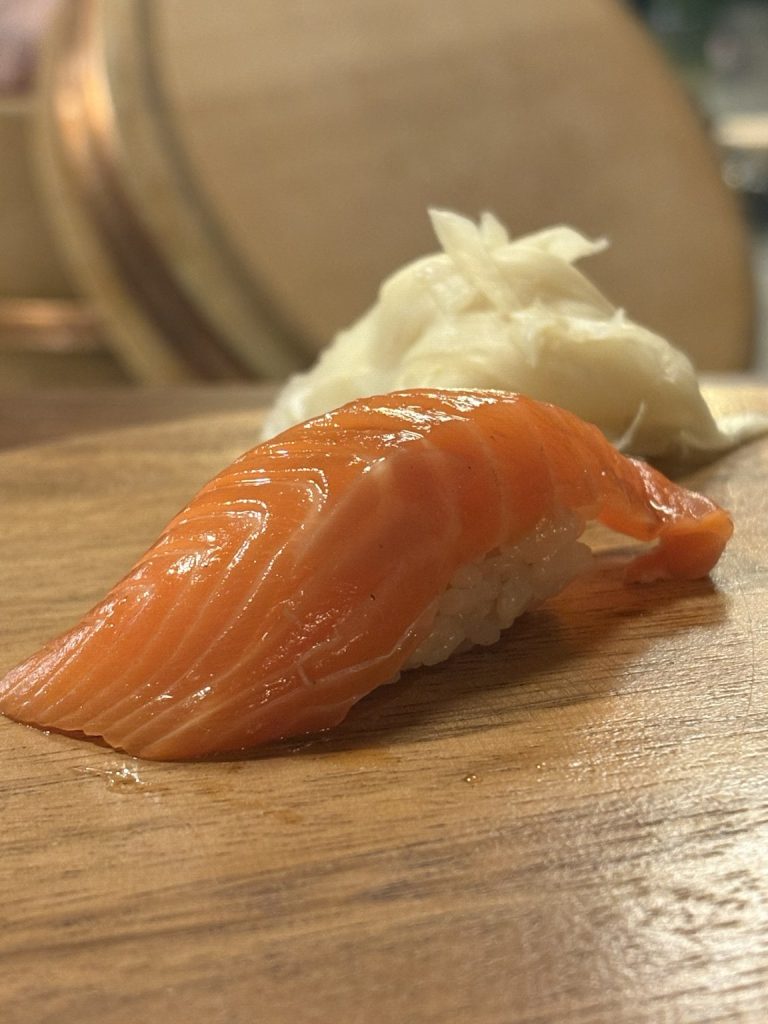 Nigiri med tjock laxskiva över risbädd på träbricka, med syltad ingefära bakom
