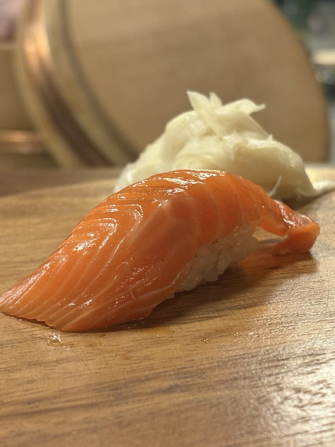 Nigiri med tjock laxskiva över risbädd på träbricka, med syltad ingefära bakom